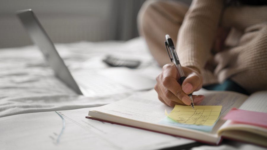A imagem mostra uma pessoa sentada na cama, com um ambiente de estudo confortável. Ela está escrevendo em um caderno, com uma caneta na mão, enquanto uma folha de post-it amarela com um esboço ou anotações é destacada. Ao fundo, um laptop e documentos espalhados indicam que o estudo está em andamento, sugerindo um ciclo produtivo de aprendizado em um ambiente informal e tranquilo.