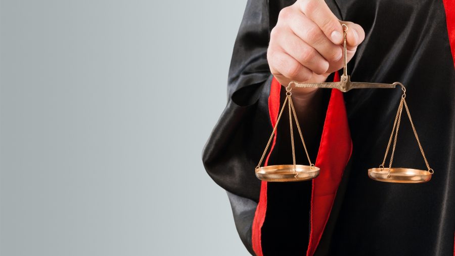 A imagem mostra uma pessoa vestindo uma toga preta com detalhes vermelhos, segurando uma balança de bronze com as duas bandejas penduradas. A balança simboliza a justiça, refletindo a imparcialidade e equilíbrio no julgamento. O fundo suave e simples ajuda a destacar o foco na balança e na vestimenta do indivíduo, sugerindo um contexto jurídico.