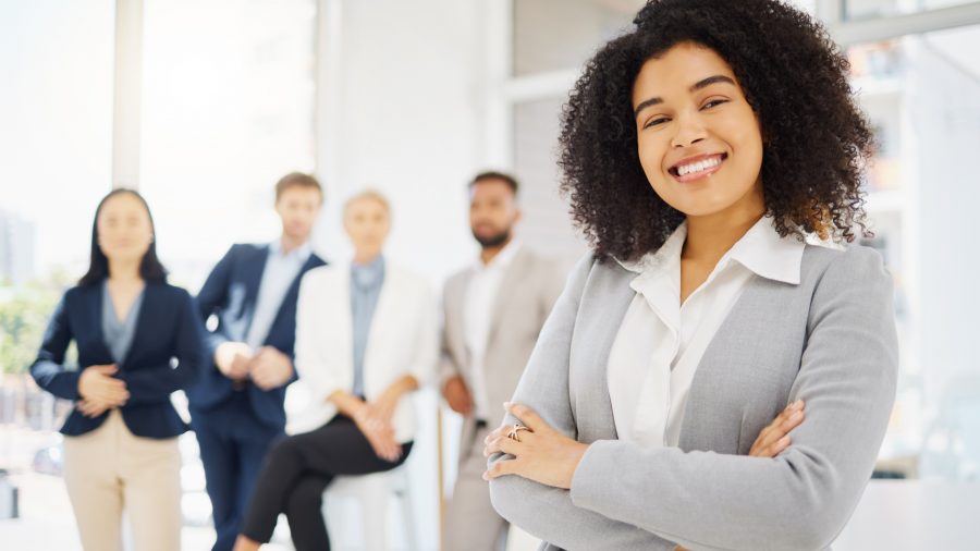 A imagem mostra uma mulher sorridente em primeiro plano, vestindo um terno cinza e com os braços cruzados, enquanto posa em um ambiente de escritório moderno. Ela está à frente de um grupo de colegas de trabalho, que aparecem desfocados ao fundo. Todos estão vestidos de forma profissional e o ambiente é bem iluminado, com janelas grandes que proporcionam luz natural. A mulher transmite confiança e postura profissional.