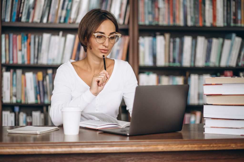 Mulher olha para notebook com expressão pensativa, segurando caneta, cercada de livros em uma biblioteca.