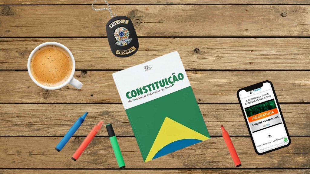 Mesa de madeira com uma xícara de café, canetas coloridas, livro da Constituição Federal do Brasil, crachá da Polícia Federal e celular aberto na página de assinatura para carreiras policiais do Decorando a Lei Seca.