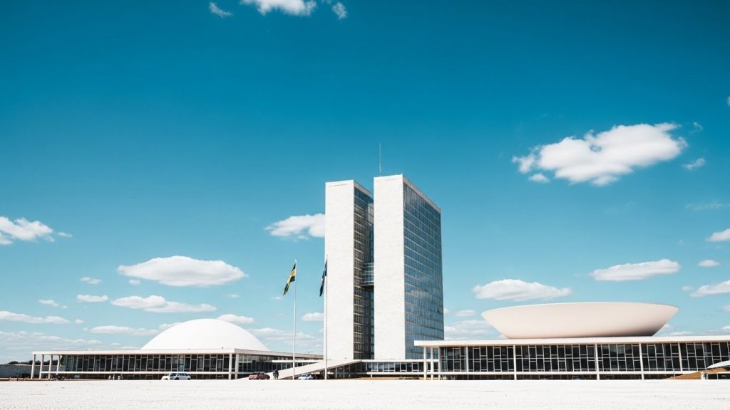 Fachada do Congresso Nacional em BrasĂlia sob cĂ©u azul com poucas nuvens, exibindo os dois edifĂcios principais: a cĂșpula branca voltada para baixo do Senado Federal, a cĂșpula voltada para cima da CĂąmara dos Deputados e as torres centrais; destaque para a arquitetura modernista de Oscar Niemeyer e bandeiras do Brasil ao centro.