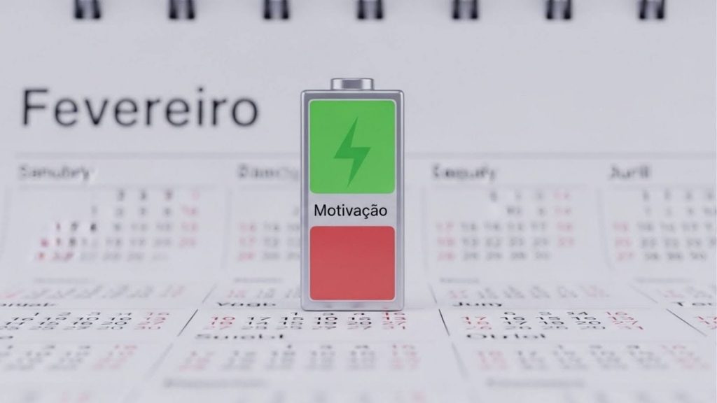 Uma bateria em 3D posicionada verticalmente no centro da imagem. A parte superior da bateria é verde com um ícone de raio, e a metade inferior é vermelha e vazia, com a palavra "Motivação" escrita no meio. Ao fundo, um calendário de mesa desfocado com o mês de "Fevereiro" em destaque.
