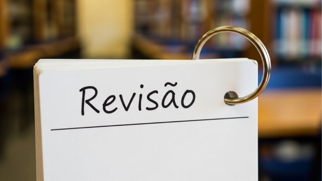 Close-up focado em um bloco de fichas de estudo (flashcards) preso por uma argola metálica prateada. Na primeira ficha branca, vê-se a palavra 'Revisão' escrita à mão em tinta preta. O fundo mostra, de forma desfocada, corredores de estantes de livros em uma biblioteca.