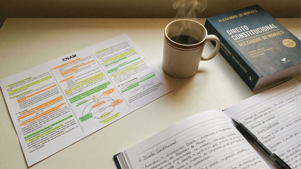 Uma mesa de estudos organizada com materiais focados em Direito Constitucional. No centro, uma folha de resumo impressa com o título "ENAM" apresenta textos destacados em marca-texto colorido e diagramas. Ao lado, uma caneca de cerâmica solta vapor, indicando uma bebida quente. À direita, um livro grosso de capa azul-marinho intitulado "Direito Constitucional - Alexandre de Moraes". Em primeiro plano, um caderno aberto exibe anotações manuscritas detalhadas com uma caneta preta sobre as páginas.