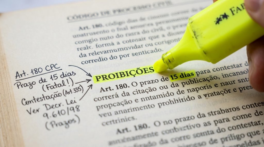 Fotografia em close-up extremo (macro) de uma página de livro jurídico impressa. Uma mão no canto direito segura um marca-texto amarelo da marca Faber-Castell e destaca a palavra "PROIBIÇÕES" e o trecho "15 dias" dentro de um parágrafo que menciona o "Art. 180". Na margem esquerda da página, há anotações manuscritas em caneta preta com setas apontando para a palavra destacada. As anotações dizem: "Art. 180 CPC", "Prazo de 15 dias (Fatal!)", "Contestação (Art. 335)" e "Ver Decr. Lei 9.610/98 (Prazos)".