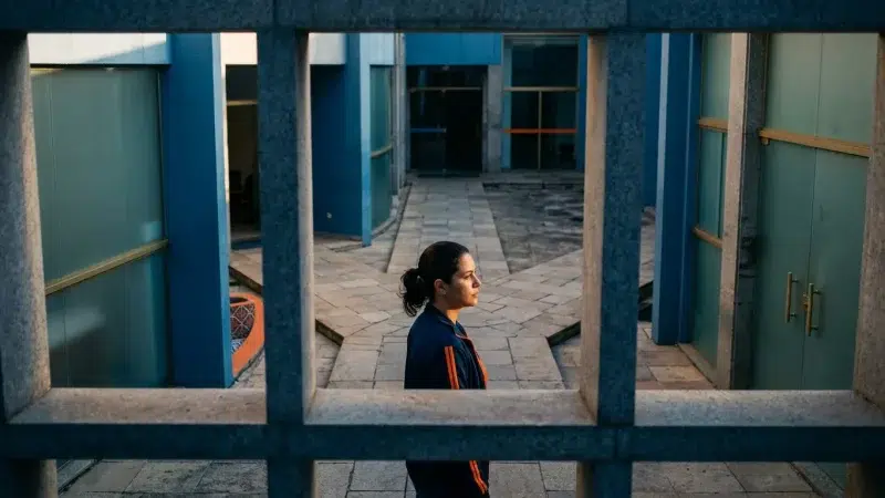 Mulher em perfil atravessando pátio de prédio público com caminhos de pedra e luz lateral