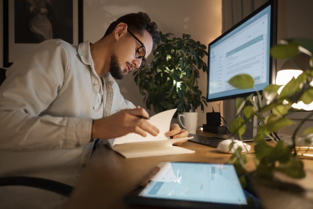 Homem usando óculos, sentado em uma mesa de trabalho organizada, estudando documentos com atenção. Na mesa, há um monitor ligado, um tablet, uma caneca e plantas ao redor, criando um ambiente aconchegante e iluminado