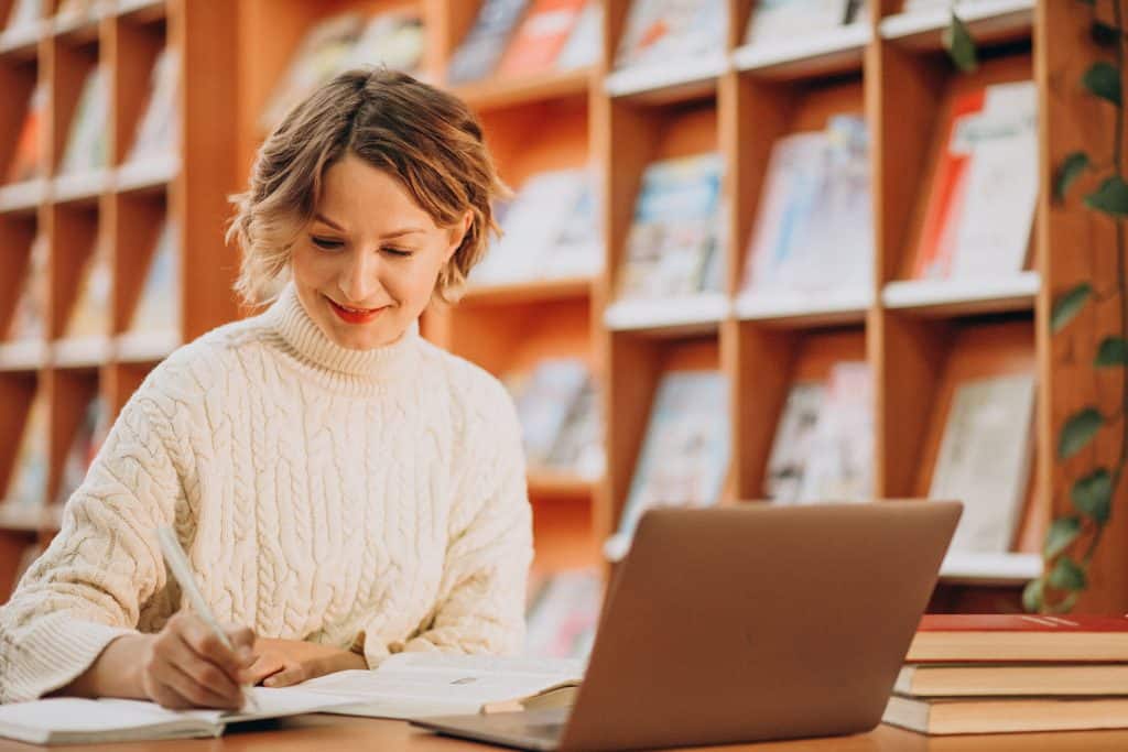 Mulher sentada em uma biblioteca, sorrindo enquanto escreve em um caderno. Ela está rodeada por livros e um laptop, com estantes de livros coloridos ao fundo, criando um ambiente acolhedor e de estudo.