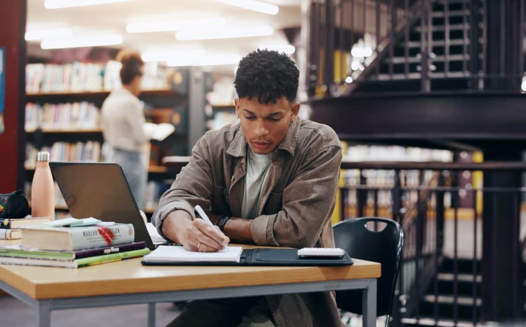 Jovem estudando em uma biblioteca, concentrado enquanto escreve em um caderno. Sobre a mesa estão livros, um laptop e materiais de estudo, sugerindo dedicação e foco em sua preparação acadêmica.