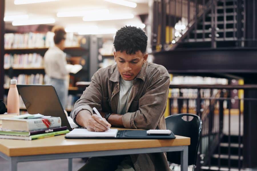 Jovem estudando em uma biblioteca, concentrado enquanto escreve em um caderno. Sobre a mesa estão livros, um laptop e materiais de estudo, sugerindo dedicação e foco em sua preparação acadêmica.