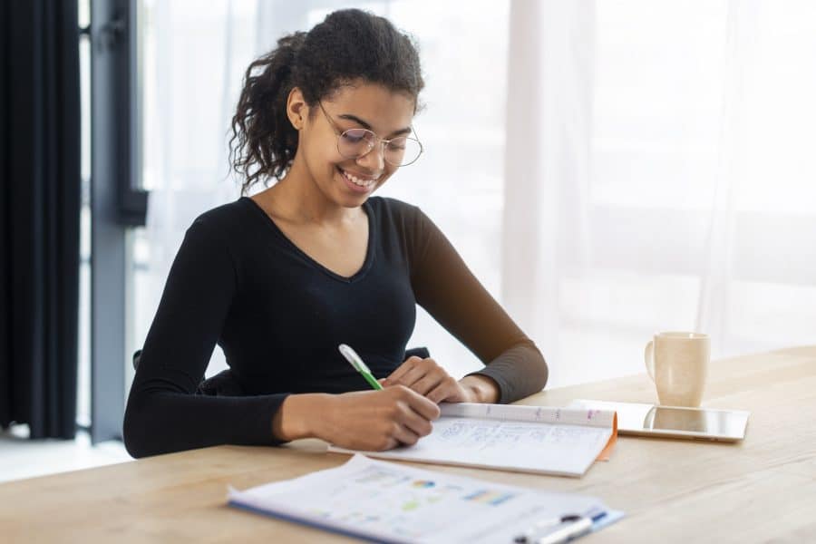 Mulher sorridente com óculos estudando em uma mesa, escrevendo em um caderno. Ao lado, há documentos e uma xícara, sugerindo um momento de concentração e dedicação, possivelmente preparando-se para provas ou concursos.