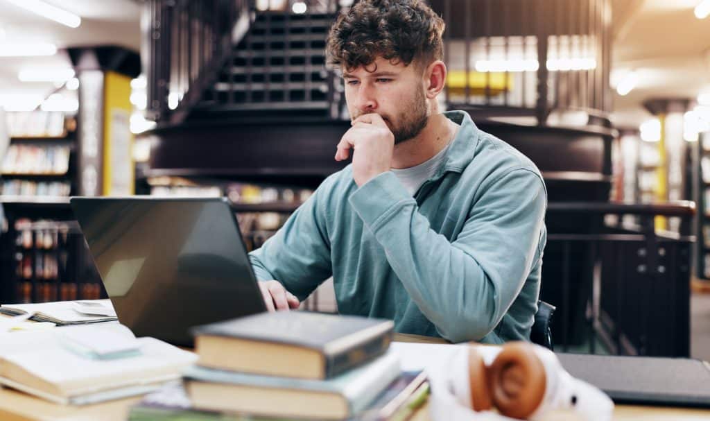 Homem concentrado estudando em uma biblioteca, com um laptop aberto à sua frente e livros espalhados sobre a mesa. O ambiente sugere dedicação e foco nos estudos, em meio a um cenário acadêmico organizado.