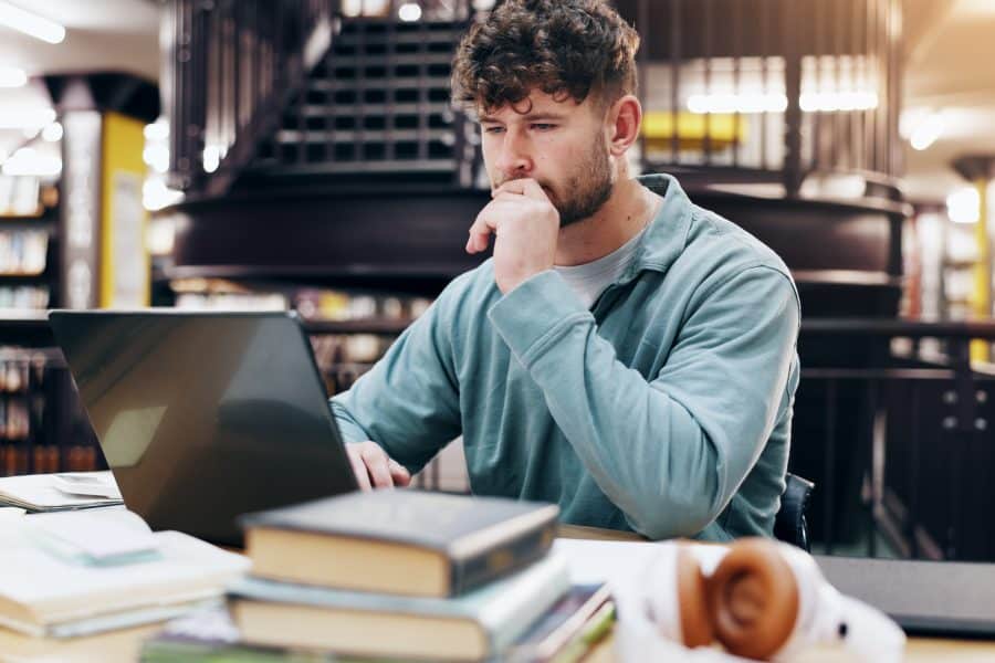 Homem concentrado estudando em uma biblioteca, com um laptop aberto à sua frente e livros espalhados sobre a mesa. O ambiente sugere dedicação e foco nos estudos, em meio a um cenário acadêmico organizado.