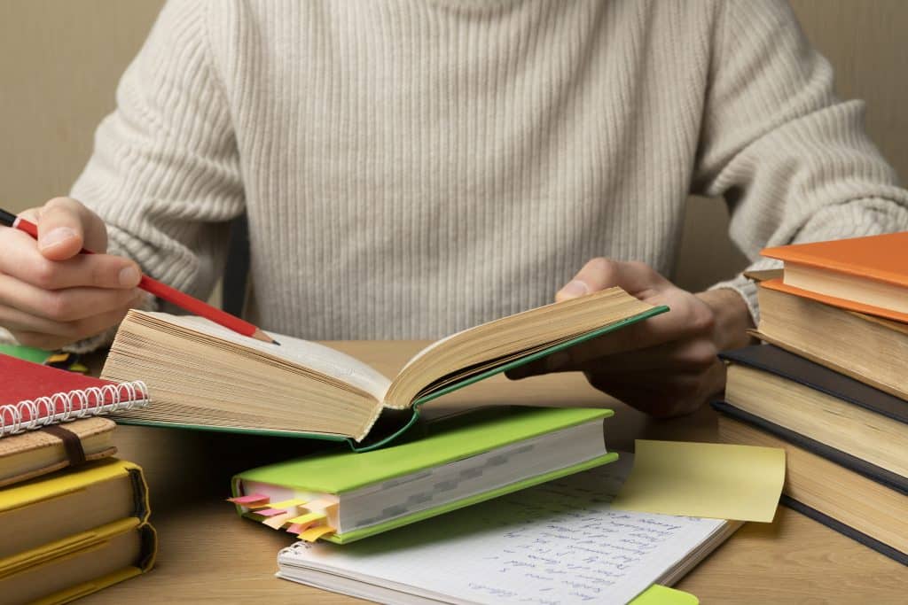 Pessoa estudando em uma mesa repleta de livros, cadernos e anotações. Com um lápis vermelho em mãos, ela consulta um livro aberto, indicando dedicação e foco na preparação para exames ou estudos acadêmicos.