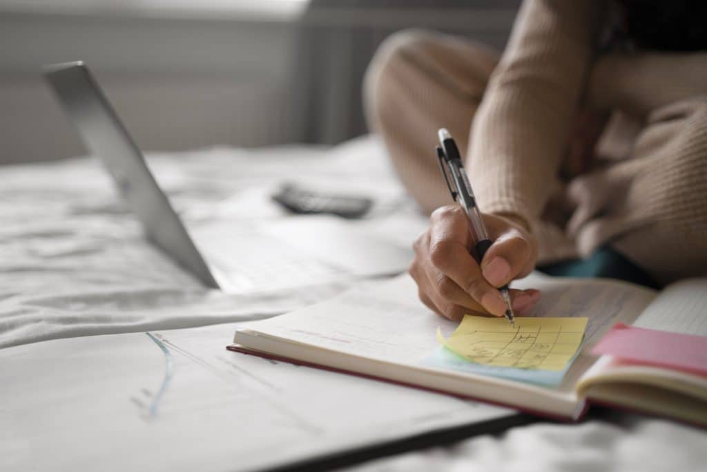 Pessoa sentada em um ambiente descontraído escrevendo em um bloco de notas adesivas sobre um caderno aberto. Ao fundo, há um laptop e documentos, sugerindo um momento de estudo ou trabalho remoto.