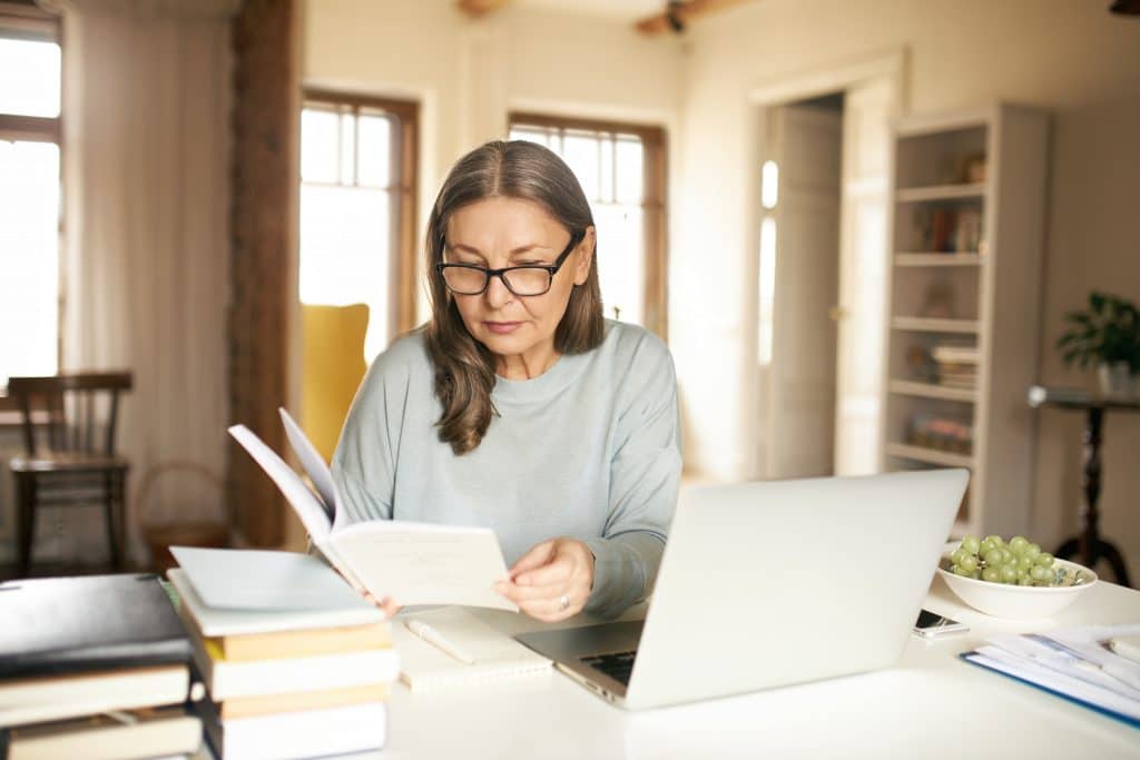 Mulher madura usando óculos, sentada em uma mesa com um laptop aberto e vários livros ao redor, enquanto lê um documento em um ambiente bem iluminado e acolhedor.