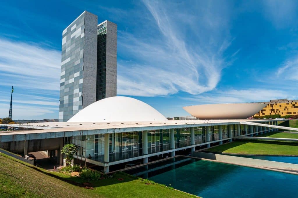 Imagem do edifício do Congresso Nacional em Brasília, sede do Poder Legislativo brasileiro, com o céu azul ao fundo e reflexo na água em primeiro plano.