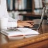 Pessoa digitando em um laptop em uma biblioteca, com livros empilhados e um caderno ao lado, sugerindo um ambiente de estudo ou pesquisa.