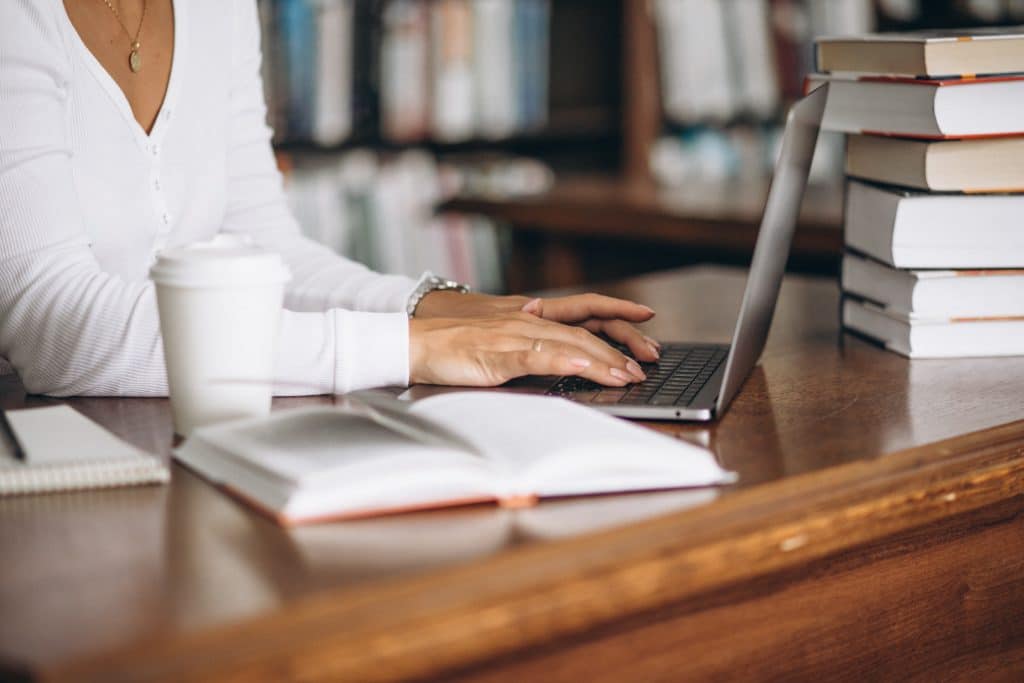 Pessoa digitando em um laptop em uma biblioteca, com livros empilhados e um caderno ao lado, sugerindo um ambiente de estudo ou pesquisa.