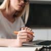 A imagem mostra uma mulher jovem, com óculos, segurando uma caneta enquanto trabalha em um laptop em um ambiente doméstico. Ela parece estar concentrada em sua tarefa, com uma folha em branco à sua frente.