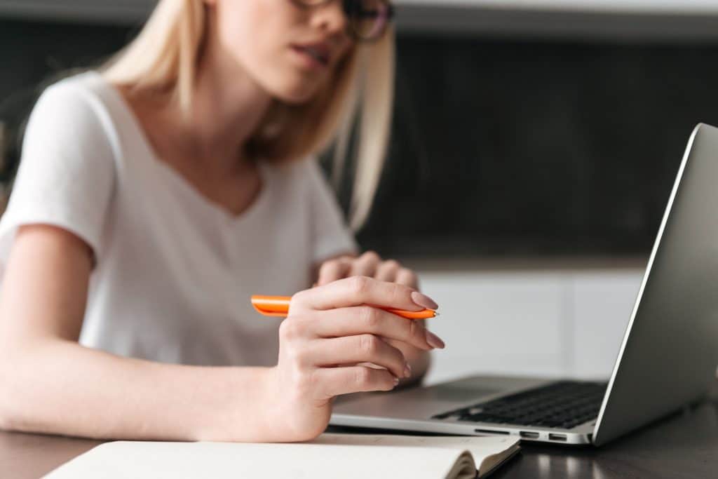 A imagem mostra uma mulher jovem, com óculos, segurando uma caneta enquanto trabalha em um laptop em um ambiente doméstico. Ela parece estar concentrada em sua tarefa, com uma folha em branco à sua frente.