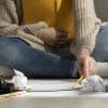 A imagem mostra uma pessoa sentada no chão, rodeada por livros, um copo de café amarelo, um caderno e papéis amassados. A pessoa está usando um lápis para escrever ou organizar o material de estudo.