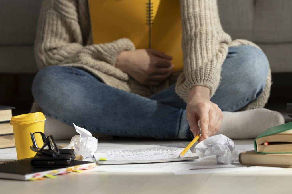 A imagem mostra uma pessoa sentada no chão, rodeada por livros, um copo de café amarelo, um caderno e papéis amassados. A pessoa está usando um lápis para escrever ou organizar o material de estudo.