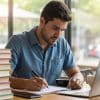 Homem sentado próximo a uma janela, ele está fazendo anotações em um caderno e com um notebook, na mesa estão diversos livros