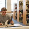 A imagem mostra um jovem concentrado lendo um livro grosso em uma biblioteca. Ele está sentado sozinho em uma mesa ampla, rodeado por estantes cheias de livros, a maioria deles técnicos ou acadêmicos. O livro que ele segura tem a palavra "LAW" (direito) na capa, indicando que o conteúdo é jurídico. O ambiente é bem iluminado, com janelas grandes ao fundo, e tem um clima calmo e organizado, ideal para estudo. A cena representa um momento de dedicação aos estudos, possivelmente de um estudante de Direito ou concurseiro se preparando para exames jurídicos.