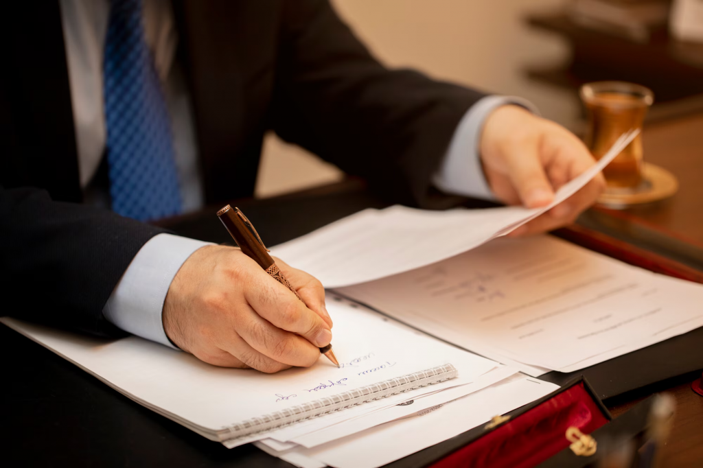 A imagem mostra um profissional, provavelmente um advogado, sentado à mesa realizando um ato jurídico. Ele está segurando uma caneta de pena e escrevendo em um papel, enquanto examina documentos que estão espalhados sobre a mesa. O cenário transmite uma atmosfera formal e focada, com a presença de um copo de bebida à direita e uma caneta de metal, sugerindo um ambiente de trabalho sério e dedicado.