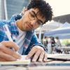 A imagem mostra um jovem estudando ao ar livre em um ambiente urbano, utilizando um caderno e um notebook sobre a mesa. Ele está concentrado, escrevendo com uma caneta enquanto consulta o conteúdo no computador. Ao fundo, há tendas e pessoas desfocadas, sugerindo que o local seja uma área pública com movimentação, como um campus universitário ou feira. A cena transmite foco, organização e conexão com o conhecimento atual.