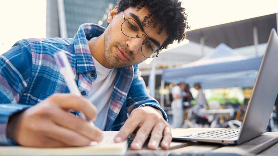 A imagem mostra um jovem estudando ao ar livre em um ambiente urbano, utilizando um caderno e um notebook sobre a mesa. Ele está concentrado, escrevendo com uma caneta enquanto consulta o conteúdo no computador. Ao fundo, há tendas e pessoas desfocadas, sugerindo que o local seja uma área pública com movimentação, como um campus universitário ou feira. A cena transmite foco, organização e conexão com o conhecimento atual.