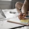 A imagem mostra uma pessoa sentada na cama, com um ambiente de estudo confortável. Ela está escrevendo em um caderno, com uma caneta na mão, enquanto uma folha de post-it amarela com um esboço ou anotações é destacada. Ao fundo, um laptop e documentos espalhados indicam que o estudo está em andamento, sugerindo um ciclo produtivo de aprendizado em um ambiente informal e tranquilo.