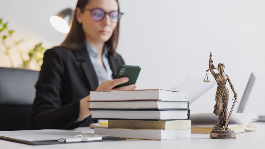 Imagem de uma mulher sentada a mesa mexendo no celular, na sua frente tem uma pilha de livros e uma figura da Justiça