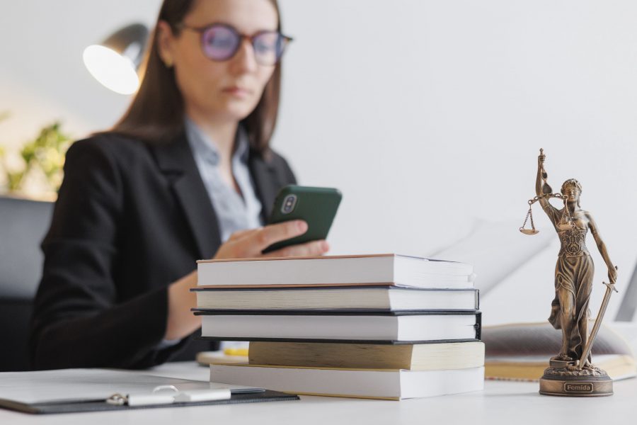 Imagem de uma mulher sentada a mesa mexendo no celular, na sua frente tem uma pilha de livros e uma figura da Justiça