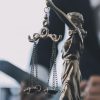 A imagem mostra uma escultura da deusa da justiça, com venda nos olhos e segurando uma balança, em destaque no primeiro plano. Ao fundo, desfocado, há uma pessoa com trajes formais manuseando documentos ou um tablet, reforçando o contexto jurídico e institucional da cena, provavelmente em um ambiente de trabalho como um escritório de advocacia ou fórum.