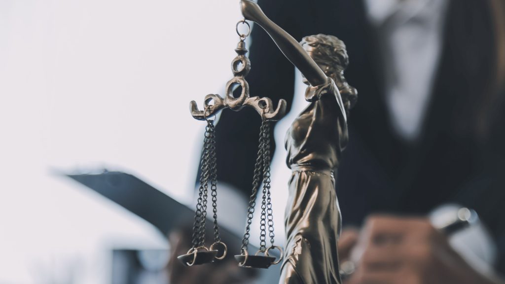 A imagem mostra uma escultura da deusa da justiça, com venda nos olhos e segurando uma balança, em destaque no primeiro plano. Ao fundo, desfocado, há uma pessoa com trajes formais manuseando documentos ou um tablet, reforçando o contexto jurídico e institucional da cena, provavelmente em um ambiente de trabalho como um escritório de advocacia ou fórum.