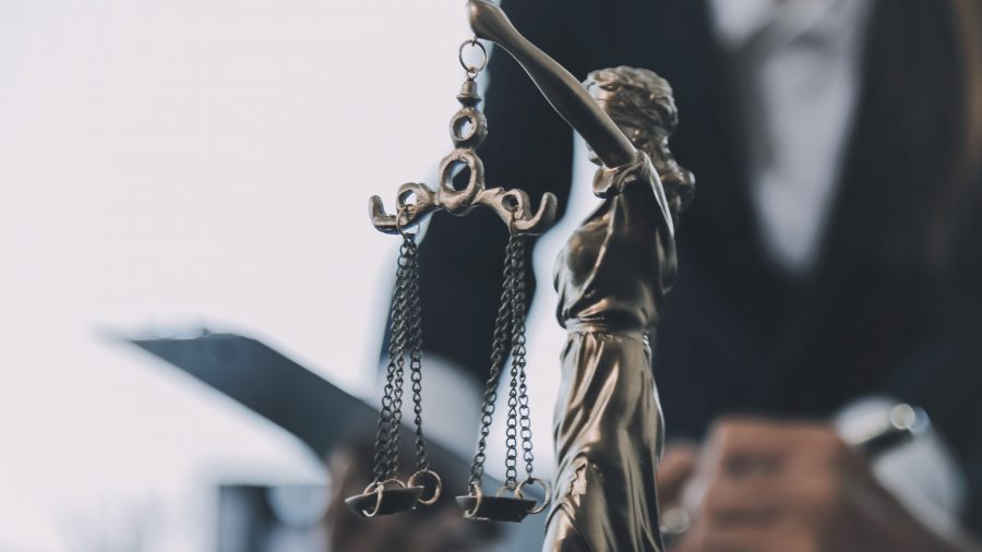 A imagem mostra uma escultura da deusa da justiça, com venda nos olhos e segurando uma balança, em destaque no primeiro plano. Ao fundo, desfocado, há uma pessoa com trajes formais manuseando documentos ou um tablet, reforçando o contexto jurídico e institucional da cena, provavelmente em um ambiente de trabalho como um escritório de advocacia ou fórum.