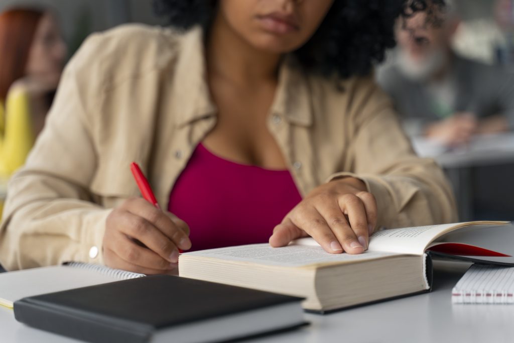 A imagem mostra uma pessoa estudando em um ambiente de aprendizado, focada em um livro aberto sobre a mesa. Ela está marcando algo no livro com uma caneta vermelha. Ao fundo, outras pessoas podem ser vistas em um ambiente descontraído de estudo, indicando uma situação de colaboração ou concentração em grupo. O cenário é claro e bem iluminado, criando uma atmosfera de estudo produtivo.