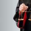 A imagem mostra uma pessoa vestindo uma toga preta com detalhes vermelhos, segurando uma balança de bronze com as duas bandejas penduradas. A balança simboliza a justiça, refletindo a imparcialidade e equilíbrio no julgamento. O fundo suave e simples ajuda a destacar o foco na balança e na vestimenta do indivíduo, sugerindo um contexto jurídico.