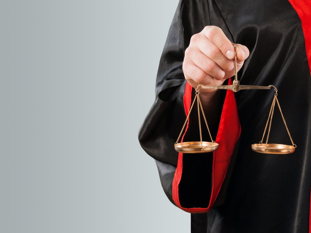 A imagem mostra uma pessoa vestindo uma toga preta com detalhes vermelhos, segurando uma balança de bronze com as duas bandejas penduradas. A balança simboliza a justiça, refletindo a imparcialidade e equilíbrio no julgamento. O fundo suave e simples ajuda a destacar o foco na balança e na vestimenta do indivíduo, sugerindo um contexto jurídico.