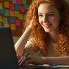 A imagem mostra uma jovem sorridente com cabelos cacheados ruivos, sentada à frente de um notebook, aparentemente estudando ou trabalhando. Ao fundo, há um quadro repleto de post-its coloridos, sugerindo uma organização visual de tarefas ou cronograma. A luz natural que entra pela janela ilumina o ambiente, criando um clima acolhedor e produtivo.