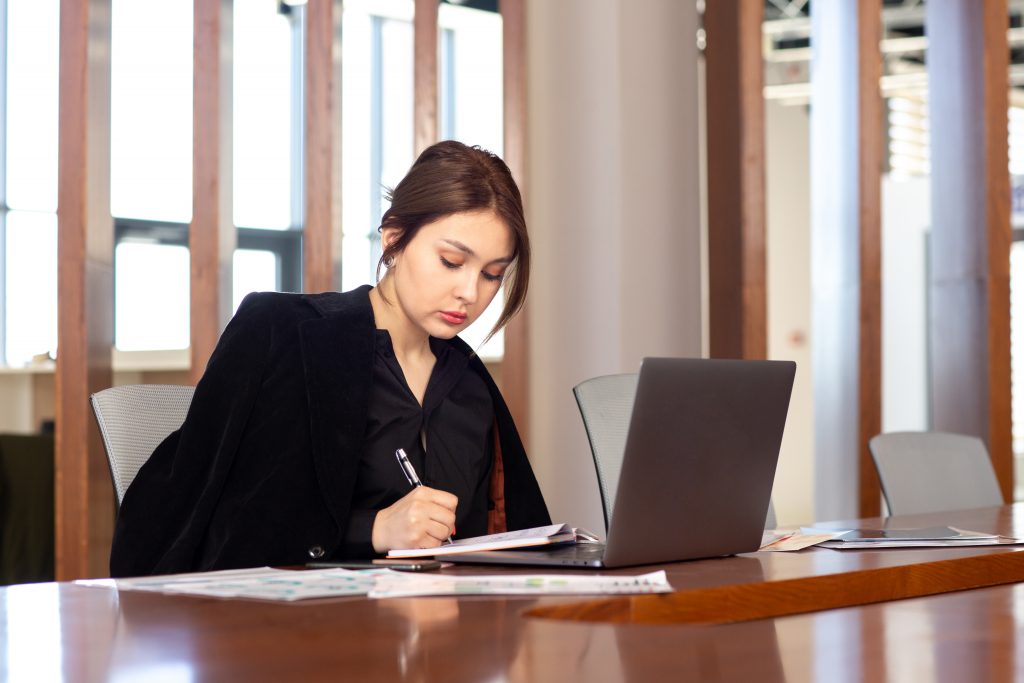 A imagem mostra uma mulher jovem em um ambiente corporativo, sentada à mesa de reunião, concentrada enquanto escreve em um caderno. À sua frente, há um notebook aberto e papéis com gráficos, sugerindo que ela está envolvida em tarefas administrativas, jurídicas ou de planejamento. Vestida de forma formal, com blazer preto, ela transmite profissionalismo e foco.