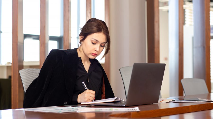 A imagem mostra uma mulher jovem em um ambiente corporativo, sentada à mesa de reunião, concentrada enquanto escreve em um caderno. À sua frente, há um notebook aberto e papéis com gráficos, sugerindo que ela está envolvida em tarefas administrativas, jurídicas ou de planejamento. Vestida de forma formal, com blazer preto, ela transmite profissionalismo e foco.