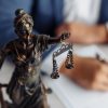A imagem mostra uma estátua da deusa da justiça, com os olhos vendados e segurando uma balança, posicionada em destaque sobre uma mesa. Ao fundo, levemente desfocado, um advogado escreve em um documento, sugerindo um ambiente jurídico, como um escritório de advocacia ou uma consultoria legal. A composição transmite seriedade, ética e compromisso com a justiça.