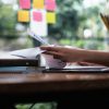 A imagem mostra uma mulher concentrada manuseando papéis e gráficos em uma mesa de estudos, cercada por cadernos, pastas e materiais de papelaria. Ao fundo, nota-se um ambiente iluminado com luz natural, janelas amplas e post-its coloridos colados no vidro, sugerindo organização visual e método ativo de estudo. O cenário transmite foco e produtividade.