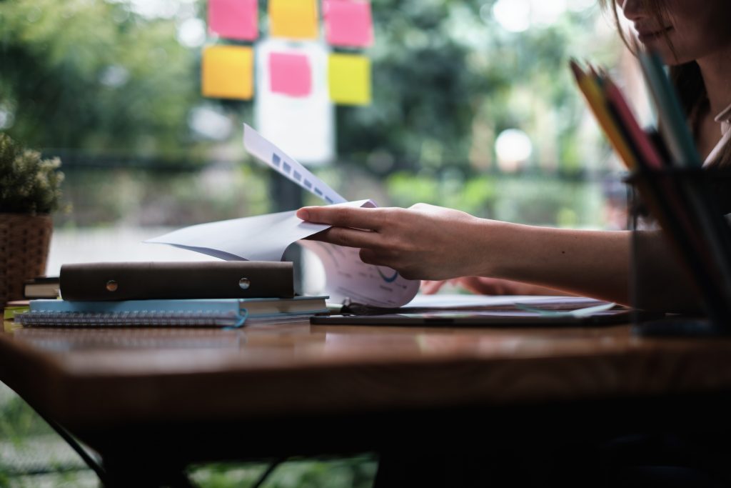 A imagem mostra uma mulher concentrada manuseando papéis e gráficos em uma mesa de estudos, cercada por cadernos, pastas e materiais de papelaria. Ao fundo, nota-se um ambiente iluminado com luz natural, janelas amplas e post-its coloridos colados no vidro, sugerindo organização visual e método ativo de estudo. O cenário transmite foco e produtividade.