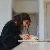 A imagem mostra uma jovem concentrada em seus estudos, sentada à mesa com um livro aberto à sua frente e um notebook ao lado. Ela segura uma caneta, demonstrando estar fazendo anotações ou marcações importantes, em um ambiente calmo e bem iluminado. A cena transmite foco, disciplina e dedicação ao aprendizado.