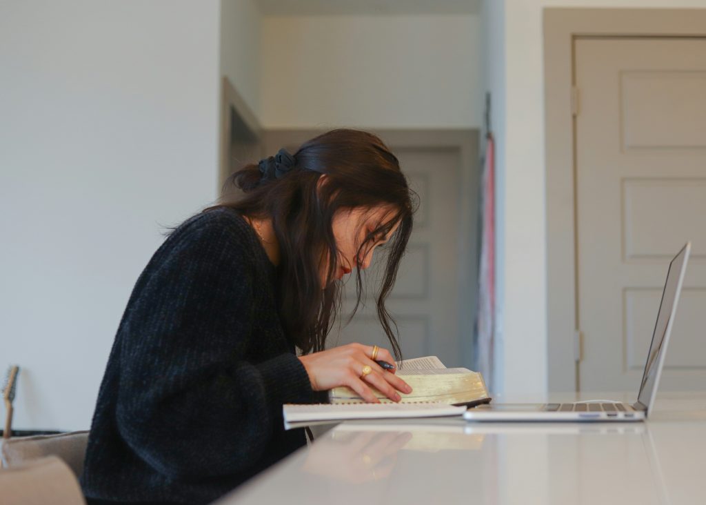 A imagem mostra uma jovem concentrada em seus estudos, sentada à mesa com um livro aberto à sua frente e um notebook ao lado. Ela segura uma caneta, demonstrando estar fazendo anotações ou marcações importantes, em um ambiente calmo e bem iluminado. A cena transmite foco, disciplina e dedicação ao aprendizado.